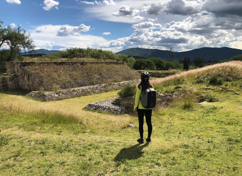 A hiker standing on a grassy field with a stone wall and mountains in the background. 4