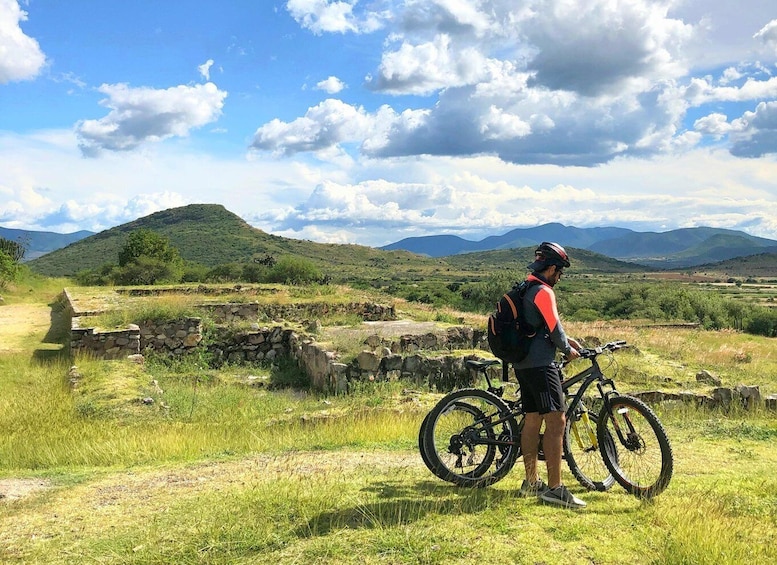 A person standing next to a mountain bike in a grassy field with stone ruins and mountains in the background. 1