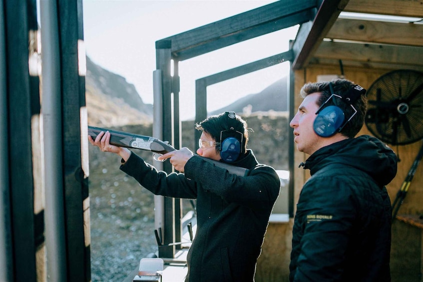 A person aiming a rifle through a window, another person observing, and a fan in the background. 1