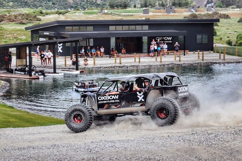 A rally car driving on a gravel path near a body of water, with spectators watching from a building. 3