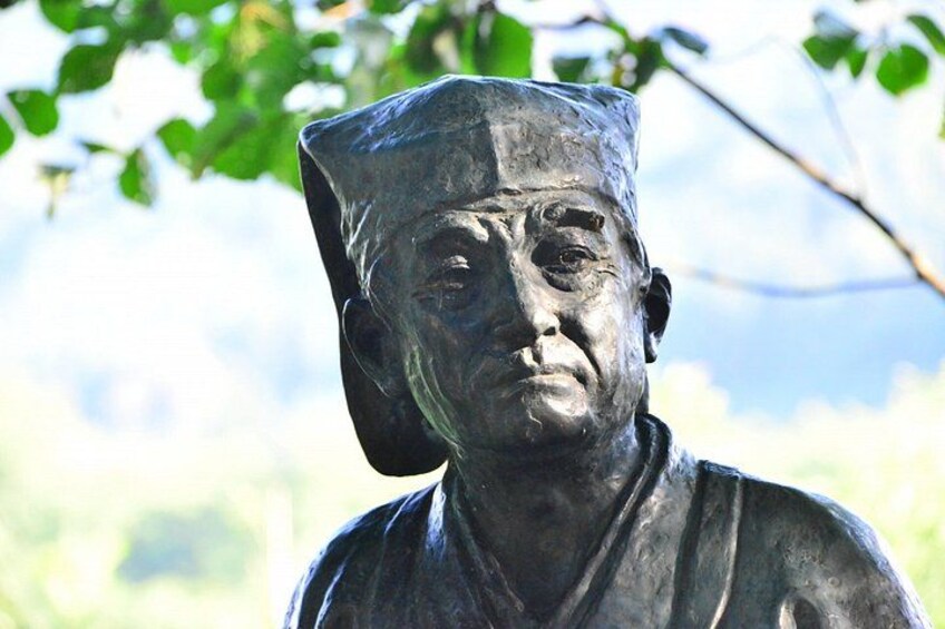A statue of a person wearing a hat, set against a blurred natural background. 3
