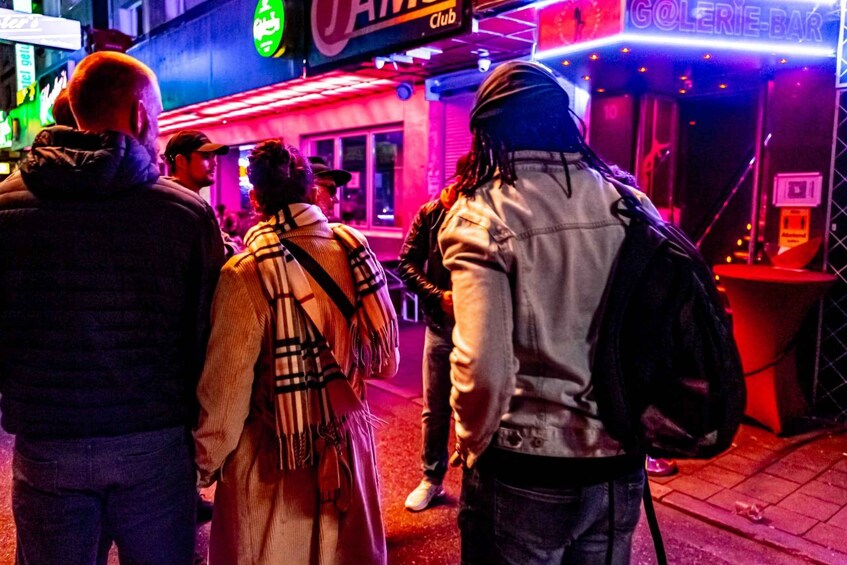 A group of people standing on a street at night with neon signs and a club in the background. 3