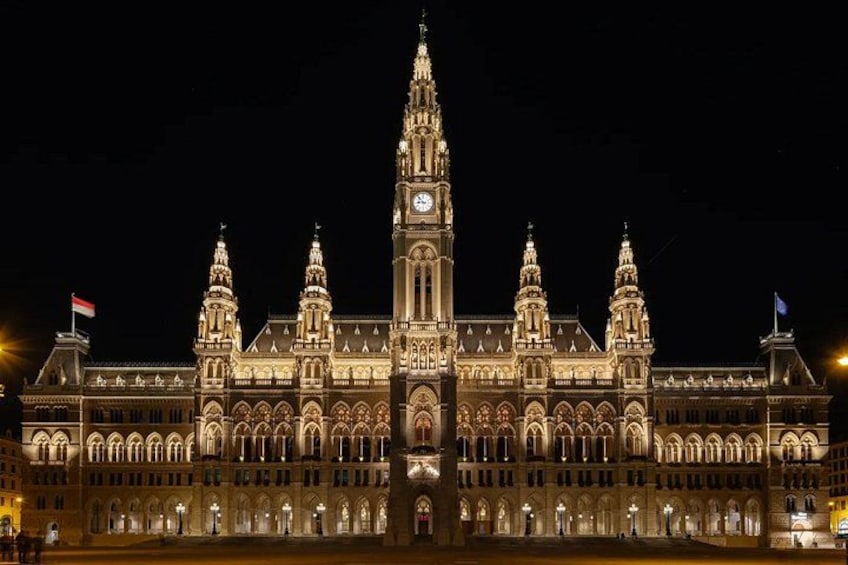 A grand, illuminated building with a central clock tower, arched windows, and flags. 2