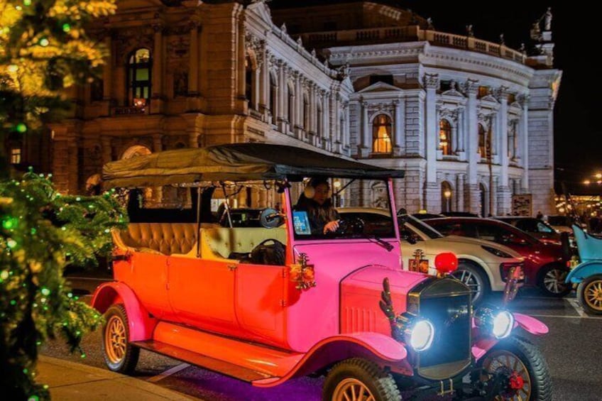 Une voiture ancienne, de couleur rose avec un toit noir, est garée devant un imposant bâtiment à l’architecture classique. 3