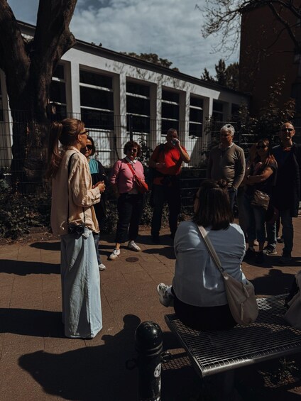 A group of people standing and sitting on a paved area with a metal bench, trees, and a building in the background. 5
