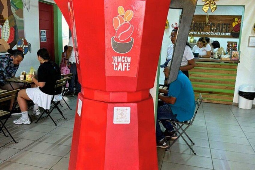 Een rode pilaar met een logo en de tekst 'Rincon Cafe', in een openbare ruimte binnenshuis waar mensen aan tafels eten. 3