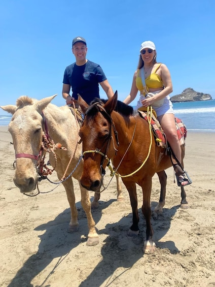 To personer rider på heste på en sandstrand med havet i baggrunden. 3