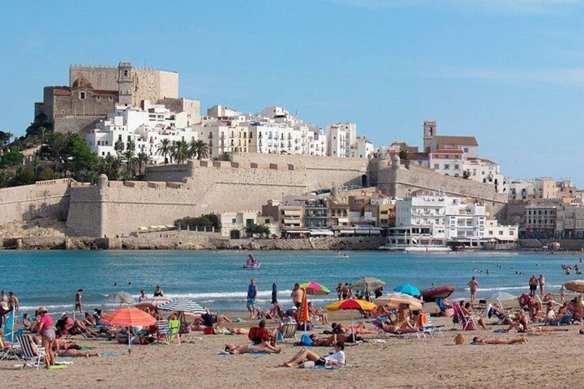 A beach with people sunbathing and swimming, a castle-like structure in the background, and a clear blue sky. 2