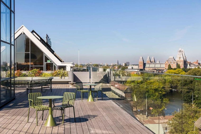 A rooftop terrace with outdoor seating, a glass building, and a view of a river and historic buildings in the background. 1