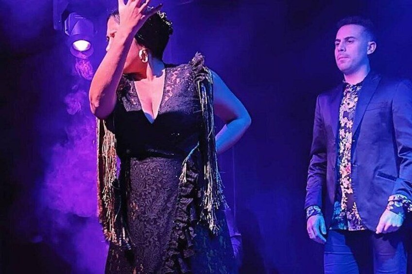 A person in a dark, patterned dress with fringe details, standing on a stage with a microphone. 3