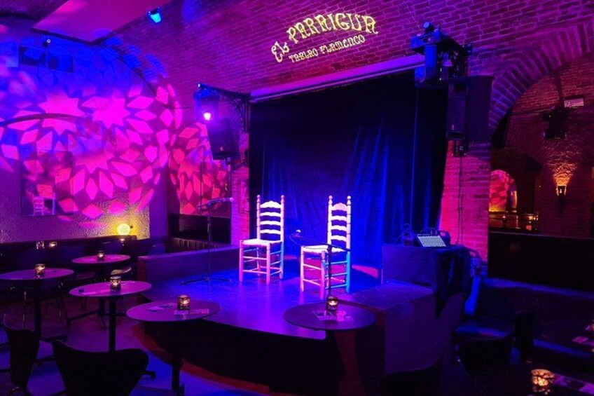 A dimly lit stage with two chairs, a brick wall, and a neon sign reading 'PERRIGUER.' 2