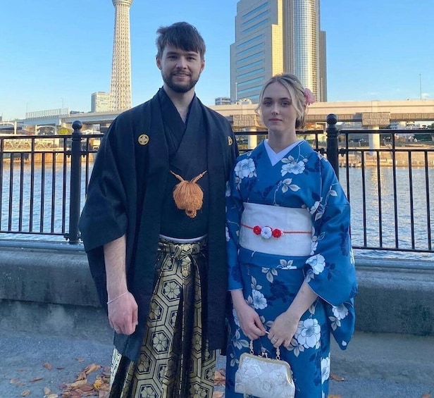 Two people in traditional Japanese attire standing by a river with modern buildings in the background. 1
