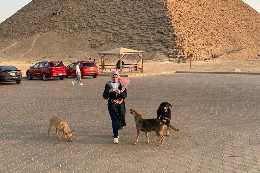 A paved area with a large pyramid in the background, a woman walking with two dogs, and parked cars. 2