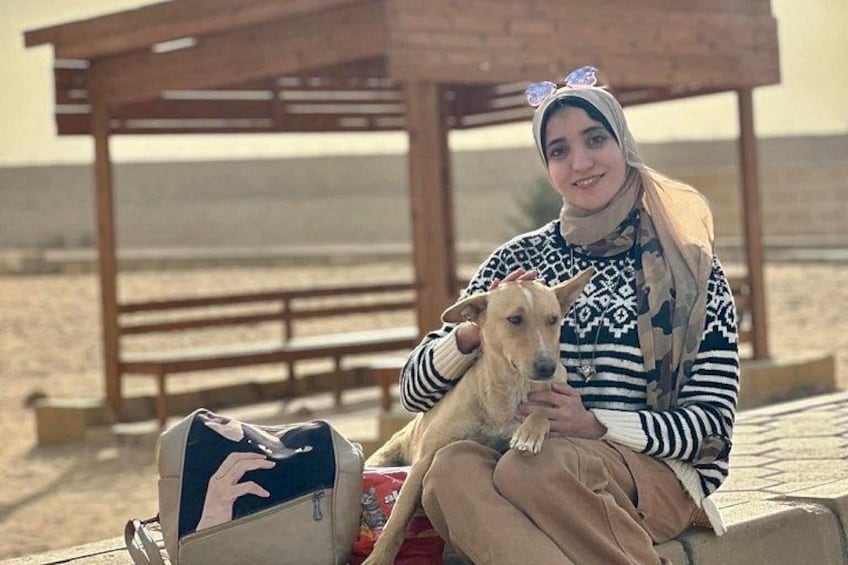 A person sitting on a bench with a dog, a wooden structure in the background, and a bag on the ground. 4