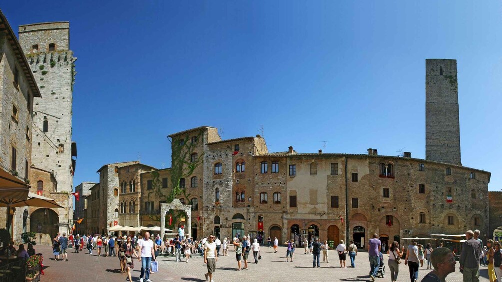 A historic square with stone buildings, a prominent tower, and a bustling crowd. 3