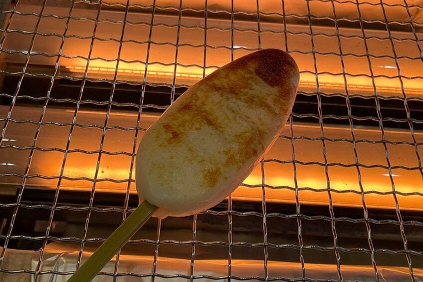 A grilled corn on the cob with a charred end, held by a green stem, against a wire mesh background. 4