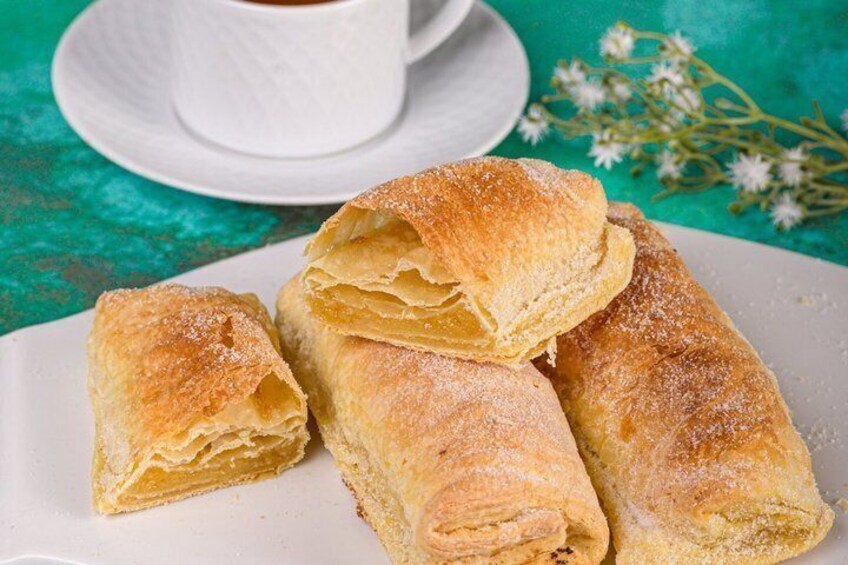 Une assiette de pâtisseries feuilletées saupoudrées de sucre glace, accompagnée d’une tasse de café sur une nappe de couleur turquoise. 3