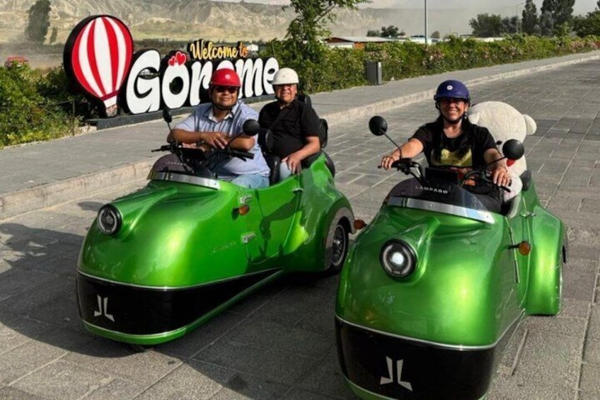 Tres personas en autos verdes antiguos, con un cartel que dice 'Welcome to Gorame' al fondo. 1