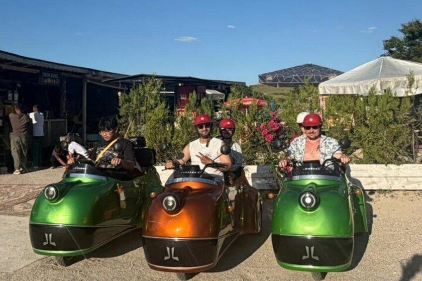 Tres personas en vehículos pequeños, verde y naranja, con 'JL' en la parte frontal, estacionados al aire libre con un edificio y árboles al fondo. 3