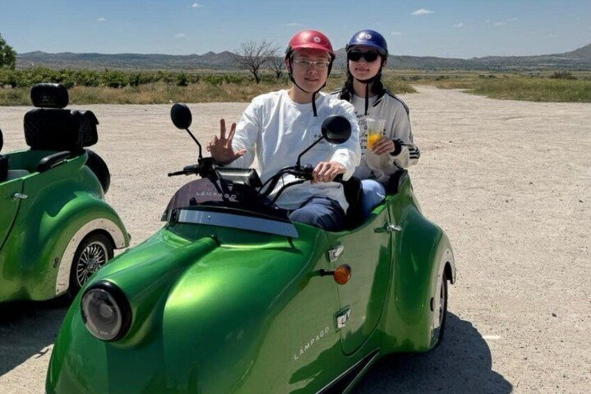 Dos personas en una motocicleta verde antigua con sidecar, estacionada en un camino de tierra con un paisaje desértico al fondo. 4