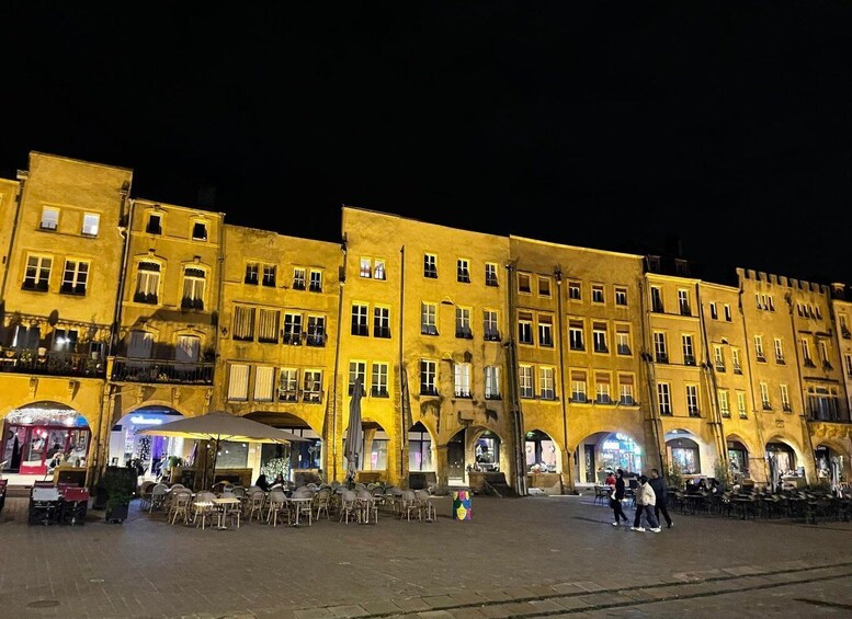 A brightly lit historic building with multiple windows and arches, a paved square with outdoor seating, and a tram in the background. 4