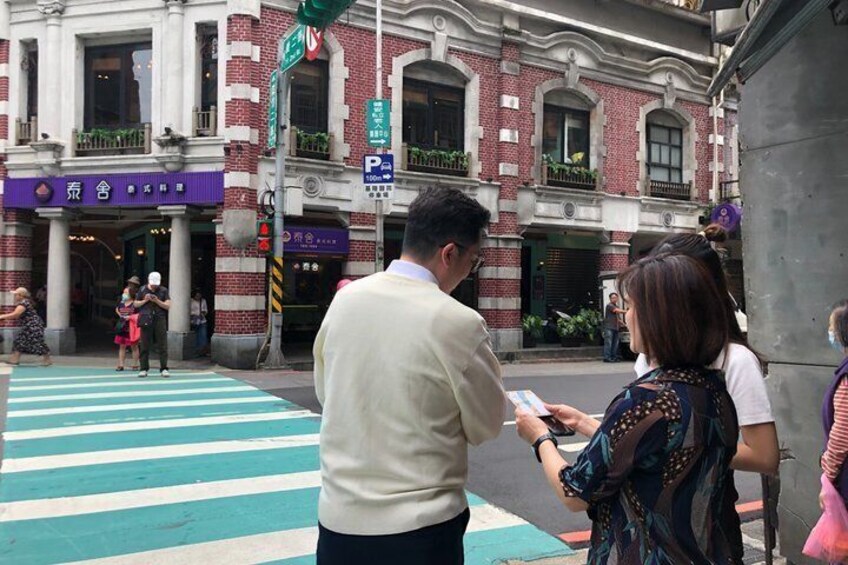 Un attraversamento pedonale con motivo a strisce, un uomo che consegna un documento a una donna, e un edificio con facciata in mattoni rossi e una insegna. 1