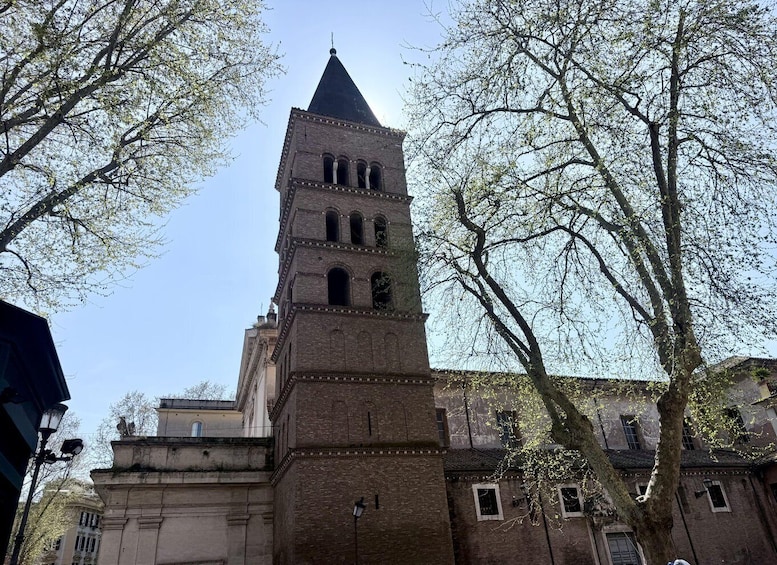 A historic tower with a pointed roof, surrounded by trees and adjacent buildings. 2