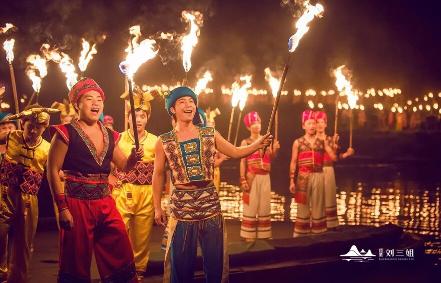 Performers in traditional costumes with torches, standing by a body of water at night. 3