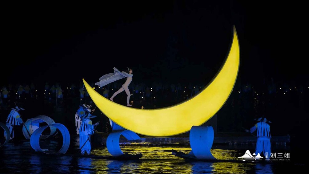 A nighttime outdoor performance with a large illuminated crescent moon structure, performers in blue costumes, and a reflective water surface. 1