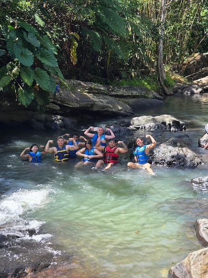 A group of people sitting on rocks in a clear stream surrounded by greenery. 3