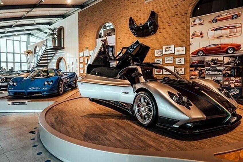 A showroom with a convertible sports car on display, a blue sports car in the background, and a brick wall with car posters. 1