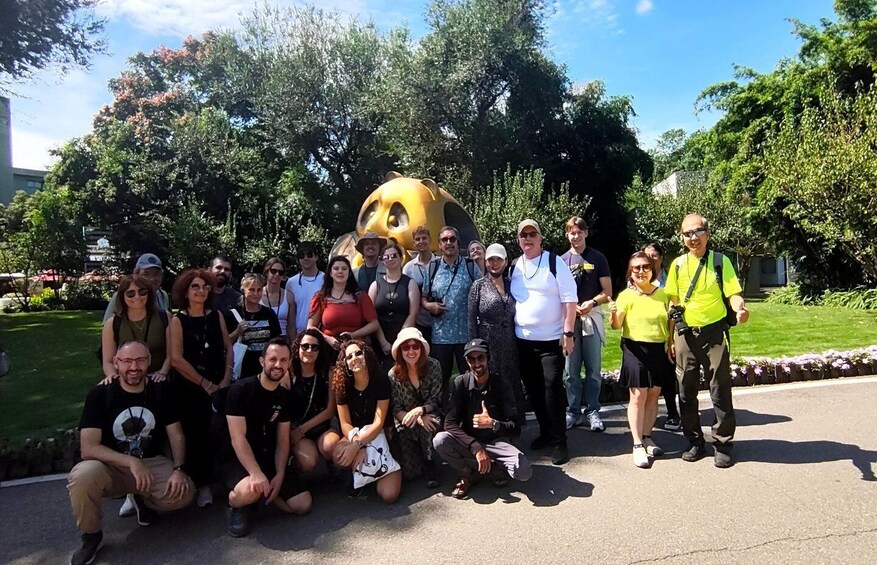 A group of people posing in a park with trees and a large yellow sculpture in the background. 1