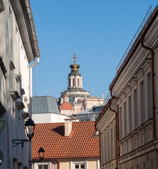 A building with a red-tiled roof and a white facade, a church with a dome and cross, and a clear blue sky. 1