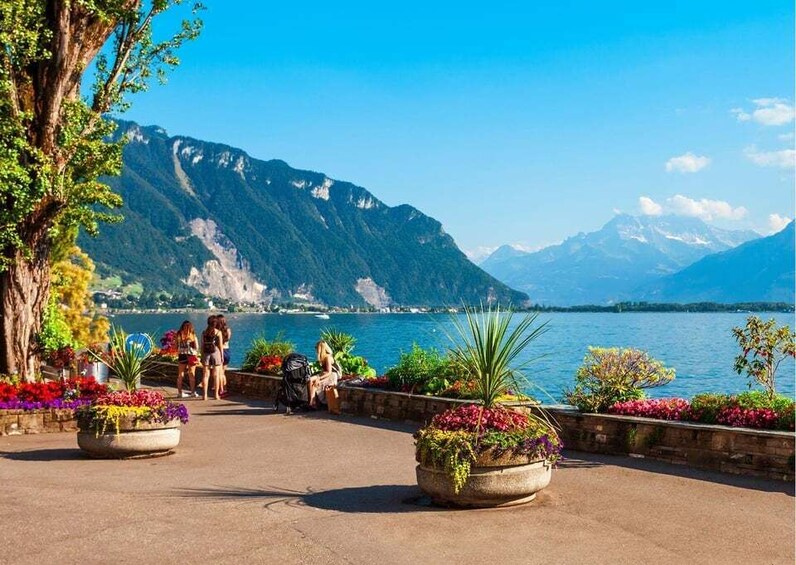 A lakeside promenade with people walking, surrounded by mountains and colorful flower beds. 4