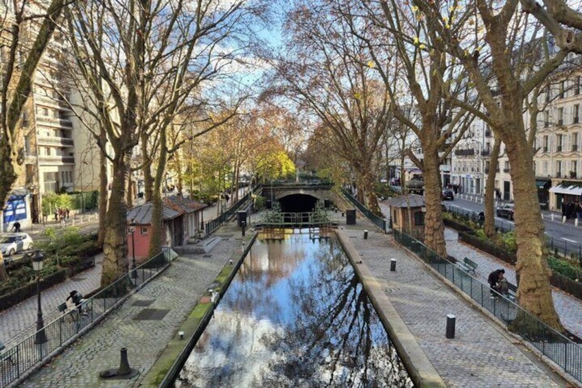 A canal with a bridge, trees lining the path, and buildings in the background. 1