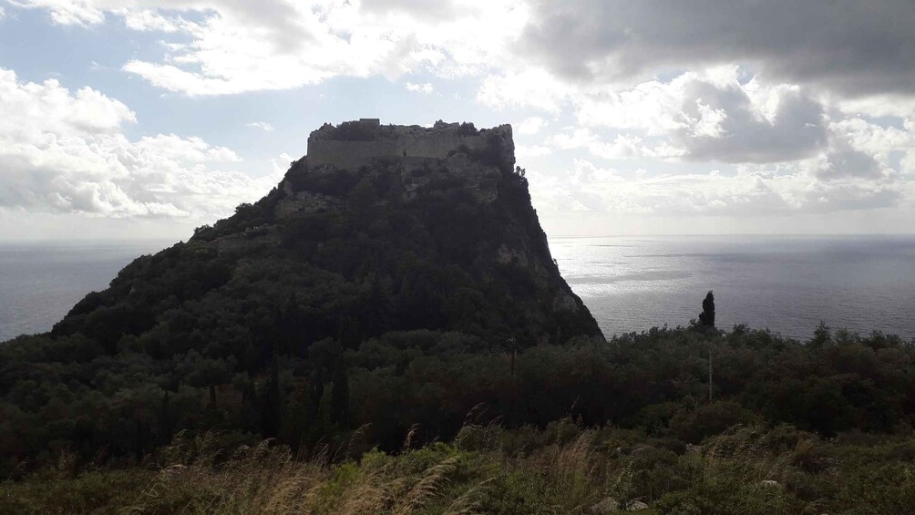 A coastal mountain with a fort on top, surrounded by dense vegetation and a view of the sea. 3