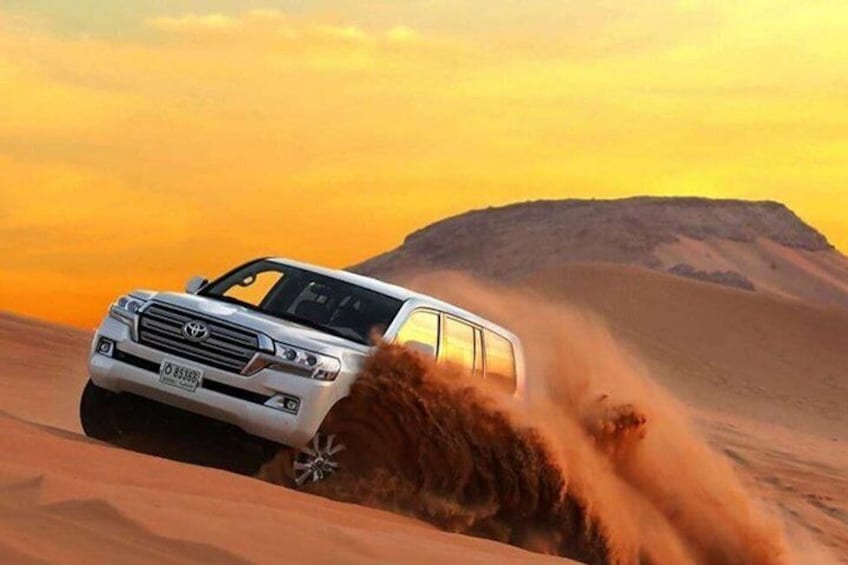 A white SUV driving through a desert with sand being kicked up, a clear sky, and a large dune in the background. 1