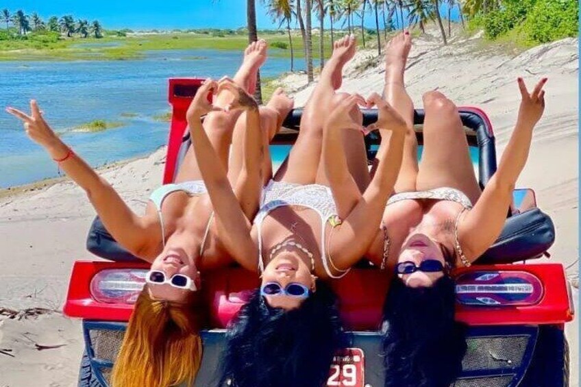 Three people lying on a red vehicle by a beach with palm trees and water in the background. 1