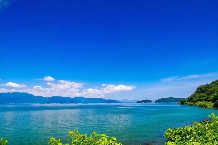 A calm lake with clear blue water, surrounded by lush greenery and distant mountains under a bright blue sky. 4