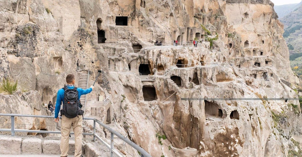 A person taking a selfie with a selfie stick in front of a rocky cliff with multiple caves. 2