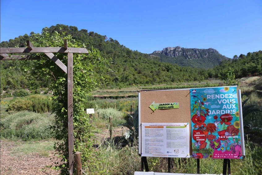 A rural landscape with a wooden pergola, a signboard with information about Jardin A Papillons, and a poster advertising an event at the gardens. 3