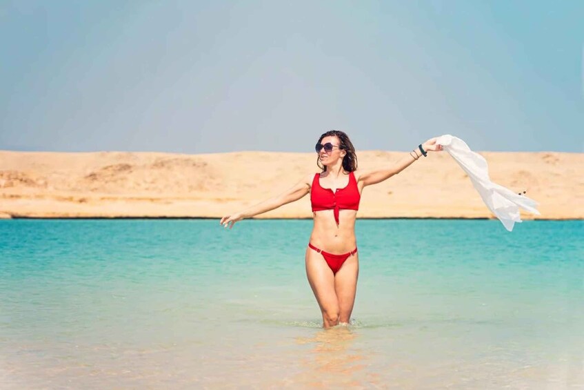 A person in a red swimsuit standing in shallow water with arms outstretched, holding a white cloth. 4