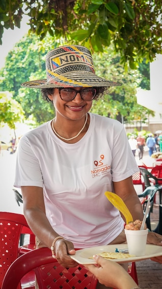 A person wearing a white t-shirt with a travel logo, seated outdoors with a hat, holding a plate with food and a drink. 5