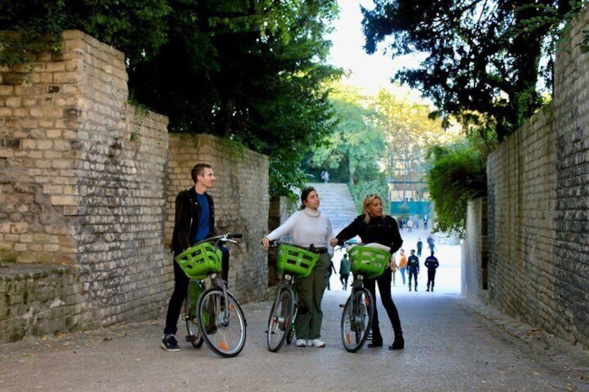 Three people on bicycles with baskets, a brick wall, and a pathway. 2
