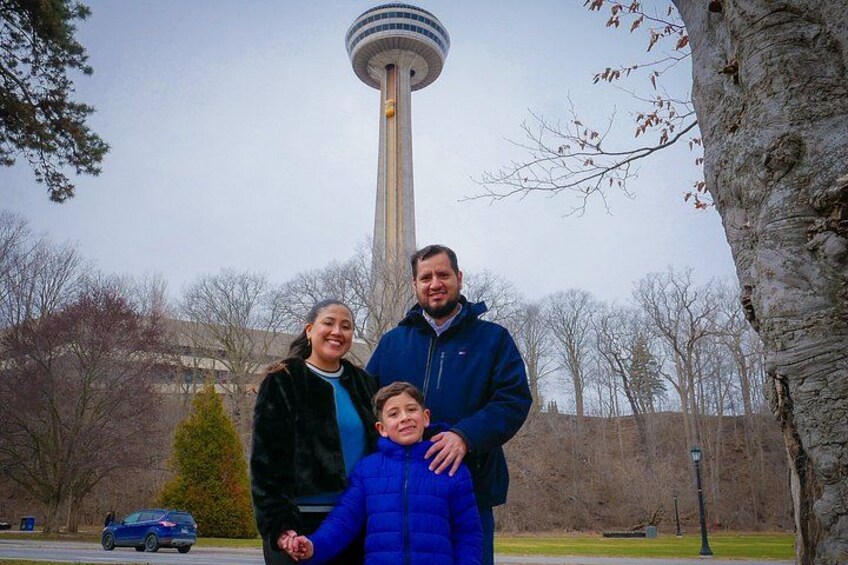 A family of three in front of a tall tower with a circular observation deck, a tree, and a parked car. 3