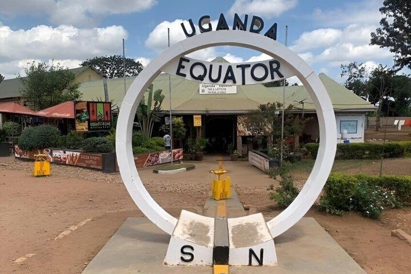 Een bord voor Uganda Equator met een ronde omlijsting, een gebouw op de achtergrond en een heldere lucht. 1
