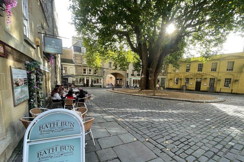 Una strada acciottolata con un caffè chiamato 'Bath Bun' e un grande albero che offre ombra. 4