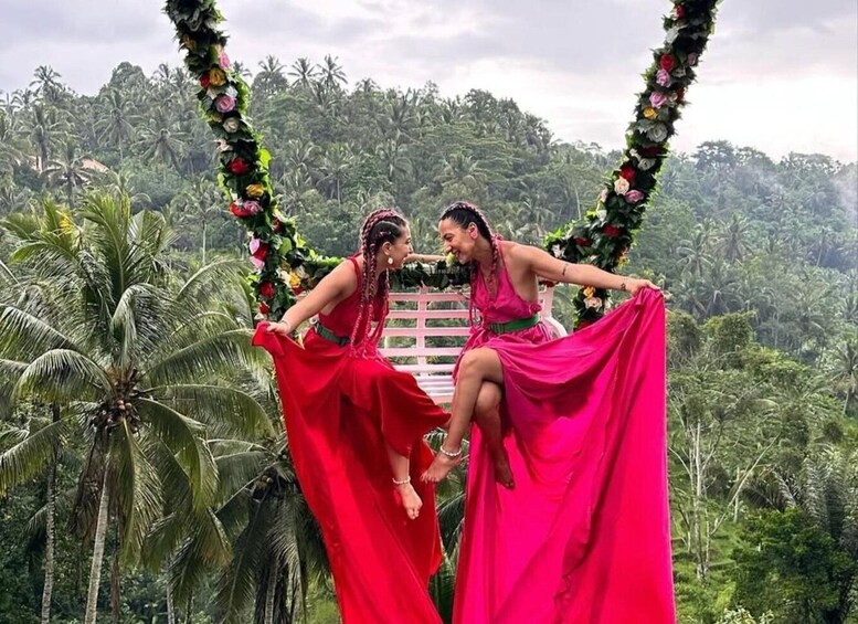 Two people in red dresses sitting on a bench with a floral arch and a lush green landscape in the background. 2