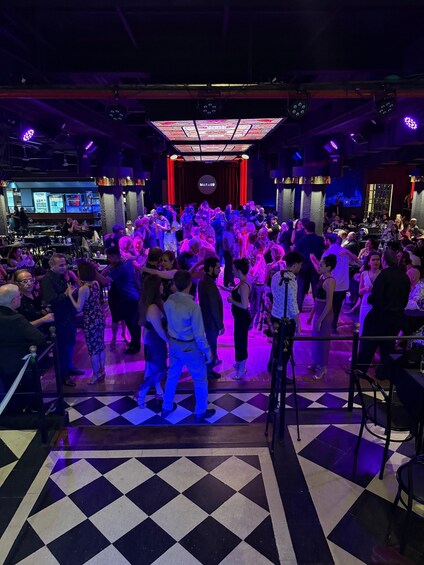 A crowded dance floor with people dancing, a checkered floor, and a stage with a red curtain in the background. 1