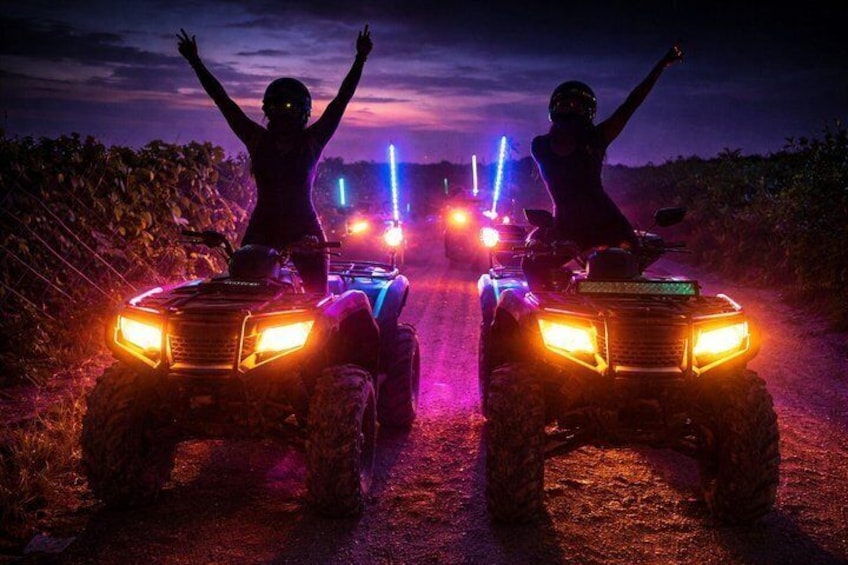 Two people on ATVs on a road with colorful lights in the background. 3